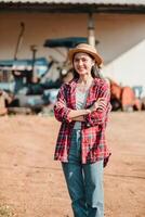 contenu agriculteur des stands avec bras franchi dans une cour de ferme, portant une paille chapeau et plaid chemise, avec agricole machinerie dans le Contexte. photo