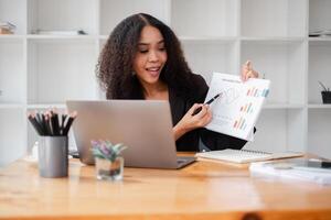 souriant femme d'affaires points à une financier graphique, engageant avec contenu sur sa portable à une meublé en bois bureau. photo