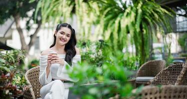 souriant avec contentement, une femme jouit sa café Pause dans une luxuriant jardin, avec sa travail posé en dehors sur le table dans de face de son. photo