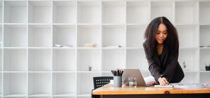 professionnel femme penche plus de sa travail bureau, intensément concentré sur l'écriture Remarques avec une portable ouvert dans de face de sa dans une moderne bureau. photo