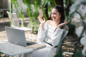 Animé et joyeux femme semble à être sur une appel, faire des gestes avec sa mains, avec une portable sur le table dans un Extérieur jardin. photo