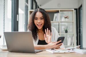 enthousiaste Jeune femme engageant dans une appel vagues Bonjour à le filtrer, avec sa portable ouvert et café sur le bureau. photo