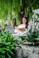 femme jouit une paisible travail réglage parmi luxuriant fougères et végétaux, l'écriture dans une carnet avec sa portable ouvert dans une serre environnement. photo