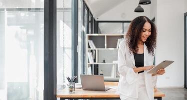 concentré femme d'affaires dans professionnel tenue est révision rapports sur sa numérique tablette, avec une portable ouvert sur le table dans une moderne bureau. photo