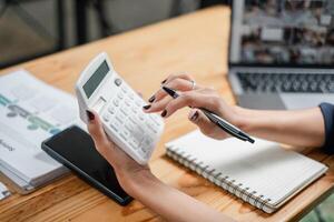 fermer de une professionnels mains en utilisant une calculatrice et prise Remarques, avec une portable et téléphone intelligent sur le bureau. photo