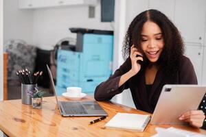 professionnel femme d'affaires engageant dans une conversation sur sa téléphone tandis que révision information sur sa tablette à sa organisé Bureau bureau. photo