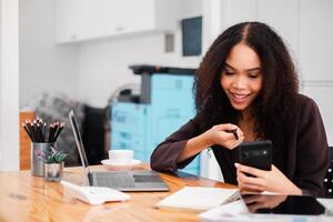 souriant femme d'affaires dans une élégant bureau, satisfait tandis que conduite une transaction sur sa téléphone intelligent, avec une portable ouvert dans de face de son. photo