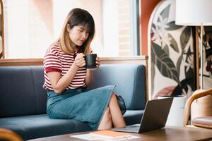 réfléchi Jeune femme en portant une café tasse, séance sur une canapé dans une confortable coin avec sa ordinateur portable, profiter une silencieux moment. photo