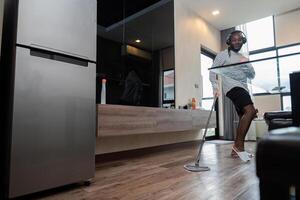 Jeune africain américain ayant amusement avec balai et la musique à maison, montrant Danse se déplace et en chantant dans vivant chambre. africain américain homme éponger en bois planchers et Faire printemps nettoyage, travaux ménagers photo