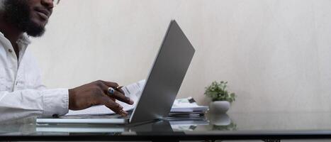 africain américain homme travail avec portable ordinateur et en utilisant calculatrice, fabrication financier Audit, révision factures impôt et comptabilité dans vivant chambre. noir gars faire free-lance travail à Accueil Bureau photo