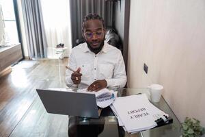 africain américain homme travail avec portable ordinateur éloigné tandis que séance à verre table dans vivant chambre. noir gars faire free-lance travail à Accueil Bureau photo