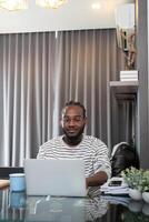 africain américain homme travail avec portable ordinateur éloigné tandis que séance à verre table dans vivant chambre. noir gars faire free-lance travail à Accueil Bureau photo