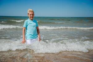 Beau content adolescent garçon de européen apparence avec blond cheveux dans blanc shorts, et une bleu T-shirt séance et éclabousser dans le mer, et regards à le caméra. été vocation concept.été Voyage vente concept.copy espace. photo