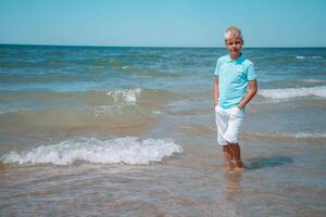 Beau adolescent garçon de européen apparence avec blond cheveux dans blanc shorts, et une bleu T-shirt des stands dans le mer dans le eau, et regards à le caméra. été vacances concept.été Voyage , famille vacances, voyage concept.copy espace. photo