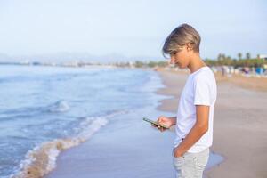 Beau adolescent garçon dans blanc T-shirt en utilisant téléphone intelligent à le plage sur été fin de semaine vacances, voyage ou voyage dans Espagne .voyage, vacances, vacances, liberté concept.côté voir. photo