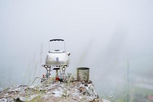 une petit bouilloire des stands sur une gaz brûleur dans le montagnes, ébullition l'eau sur une touristique brûleur, boisson thé sur une randonnée, Montagne randonnée, camping ustensiles, brouillard pierre photo