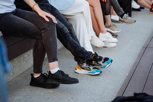 gens du repos séance sur le rue de le ville, une personne pieds dans une ligne, baskets de différent couleurs, rue vêtements, des chaussures pour le ville, noir jeans, décontractée style. photo