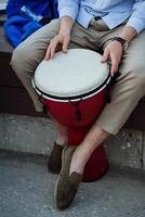 un Bureau ouvrier choisi en haut une tambouriner, une gars dans pantalon et baskets séance sur une banc en jouant une djemba, un africain tambouriner, une musical percussion instrument. photo