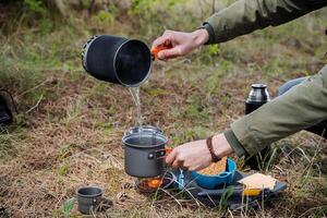 une touristique verse chaud l'eau de une pot dans une pot, cuisiniers nourriture dans le camp, petit déjeuner sur une une randonnée par le forêt, brasseries nourriture avec chaud eau, verse liquide. Humain mains photo