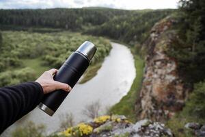 une thermos dans le main de une la personne contre le Contexte de une rivière, randonnée dans le montagnes, une paysage de montagneux terrain dans Russie, le du sud Oural, le tour de le rivière plier. photo