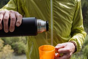 été vacances dans nature, une homme verse thé dans une verre de noir thermos, le déjeuner dans la nature sur une une randonnée par le forêt, une voyageur repose. photo