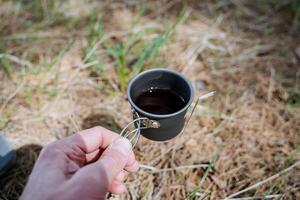 une petit agresser pour métal thé, une verre avec poignées, une compact Taille de camping ustensiles, une main en portant une tasse de café, boisson une chaud boire. photo