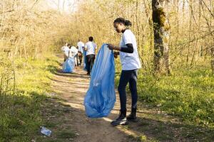 africain américain fille collecte des ordures dans une disposition bleu sac, justificatif la nature préservation et portion protéger le environnement. fier adolescent faire du bénévolat à choisir en haut poubelle. photo