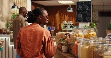 africain américain client examine bio fait maison masse des produits, à la recherche à acheter Naturel éthiquement source nourriture alternatives pour en bonne santé nutrition. femme achats pour des biens à zéro déchets magasin. caméra 2. photo