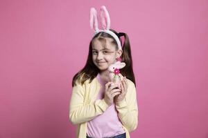 positif de bonne humeur fille avec lapin oreilles posant plus de rose arrière-plan, sentiment enthousiaste à propos Pâques de fête fête. mignonne petit enfant en portant une rose lapin jouet dans de face de caméra, duveteux articles. photo