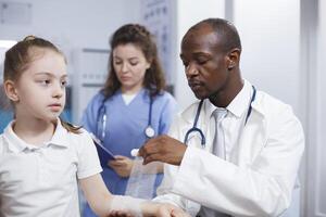 fermer de une patient bras blessure étant méthodiquement enveloppé dans stérile bandage par multiculturel médical ouvriers. Jeune enfant est étant traité par une médecin et une infirmière dans cette photographier. photo