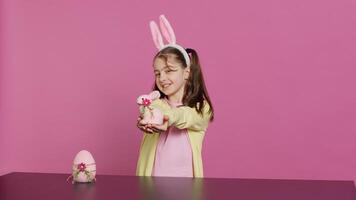 souriant bambin avec lapin oreilles montrant sa Fait main Pâques ornements, en présentant une fabriqués à la main décoré Oeuf et lapin. Jeune sucré écolière en portant de fête décorations. caméra un. photo