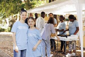 portrait coup de une mère et fille, justificatif faim le soulagement initiative à un Extérieur nourriture banque. deux caucasien femelles portant bleu t-shirts écrit bénévole, prêt à Aidez-moi le sans abri personnes. photo