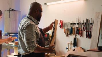 africain américain homme dans travail du bois magasin inspecter bois pièce avant assemblage meubles, vérification pour rayures. ébéniste évalue Charpente bloc, assurer il se rencontre qualité normes, caméra une photo