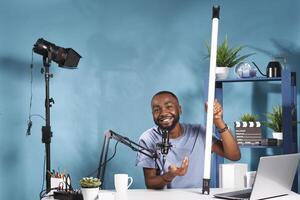 souriant blogueur en portant tube lumière équipement et expliquant Caractéristiques tandis que enregistrement pour en ligne canaliser. vlogger montrant LED lampe pour la photographie studio et vivre diffusion produit la revue photo