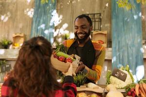 souriant africain américain vendeur remise plus de variété de biologique saisonnier produire à caucasien femelle client. de bonne humeur petit ferme affaires propriétaire avec tablier donnant boîte plein de Frais des fruits et légumes. photo