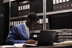 africain américain homme d'affaire en cours d'analyse administratif des dossiers, en train de lire comptabilité rapport dans entreprise dépôt. comptable travail en retard à nuit à bureaucratie record dans espace de rangement pièce photo
