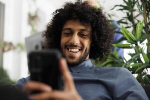 arabe homme d'affaire navigation l'Internet sur téléphone intelligent tandis que relaxant dans début en haut affaires bureau. souriant Jeune entreprise employé défilement social médias sur mobile téléphone à travail photo