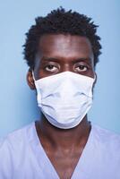 fermer de médical assistant avec visage masque à la recherche à caméra dans une studio. portrait de africain américain homme travail comme infirmière, portant bleu uniforme et ayant protection contre convoitise 19. photo