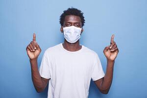 portrait de noir homme portant masque pour coronavirus protection est montrer du doigt en haut avec le sien deux indice des doigts. africain américain gars portant visage masque tandis que Faire signe avec mains dans le studio. photo