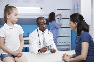 amical médecin examine enfant dans le hôpital Bureau tandis que en portant un radiographie. africain américain homme est une médical spécialiste qui fournit soins de santé prestations de service tel comme traitement et évaluation. photo