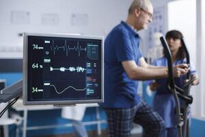 fermer de cœur taux moniteur prise une impulsion de Sénior patient pendant physique réhabilitation activité. écran montrant vital panneaux de personnes âgées homme tandis que il les usages Stationnaire vélo pour physiothérapie. photo