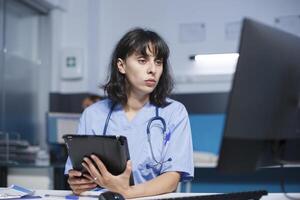 caucasien infirmière dans médical uniforme en cours d'analyse ordonnances pour les patients sur tablette et ordinateur, travail dans une hôpital bureau. médecin assistance comparant soins de santé Les données sur numérique dispositifs. photo