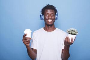 portrait de content africain américain homme permanent dans de face de une bleu Contexte avec une mis en pot plante tandis que porter une boisson. souriant noir gars en portant une café tasse et portant sans fil écouteurs. photo