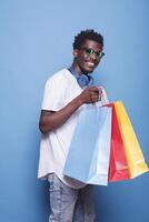 africain américain gars portant des lunettes de soleil, en portant achats Sacs, et permanent dans de face de un isolé bleu Contexte. noir homme avec sans fil casque sur le sien cou, porter coloré colis. photo