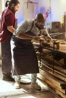 africain américain fabricant sculpture complexe dessins dans bois en utilisant ciseau et marteau, portant sécurité lunettes. artisan dans travail du bois atelier façonner en bois pièces en utilisant protecteur équipement photo