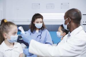 dans une clinique, noir médecin examine femelle enfant et communique avec le mère à propos santé. visage masque porté, thermomètre utilisé où multiracial médical Personnel souligner soins de santé et médicament. photo