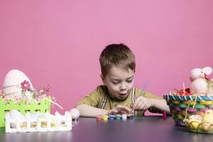 adorable bambin sourit comme il les usages aquarelles et des peintures à décorer des œufs et centres de table pour le Pâques fête festivité. peu en extase Jeune garçon explorant la créativité avec art fournitures. photo