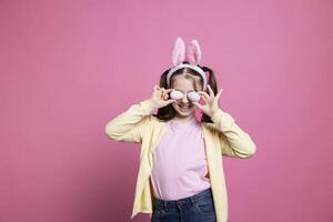 de bonne humeur enfant avec lapin oreilles tromper autour dans studio, en portant Fait main peint Pâques des œufs à célébrer avril festivité. bambin avec nattes posant avec confiance dans de face de caméra. photo