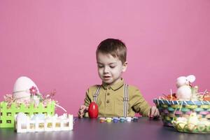 charmant petit garçon décorer des œufs avec aquarelles et empreintes tandis que fabrication magnifique ornements pour Pâques dimanche événement. adorable enfant jouit coloration avec artistique Provisions dans le studio. photo