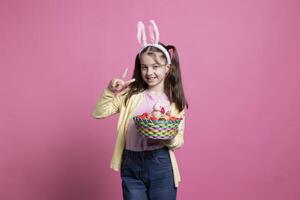 peu fille vitrines Pâques esprit par en portant charmant jouets dans le studio, montrant paix signe dans de face de caméra. petit enfant avec lapin oreilles et nattes posant avec des œufs panier et une lapin. photo